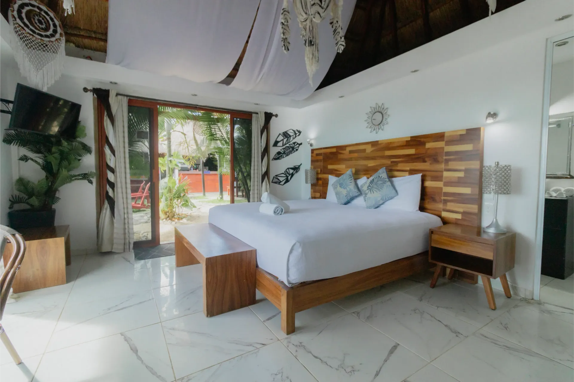 Habitación luminosa de estilo tropical con cama de madera, ropa de cama blanca y puertas que dan a un patio soleado.