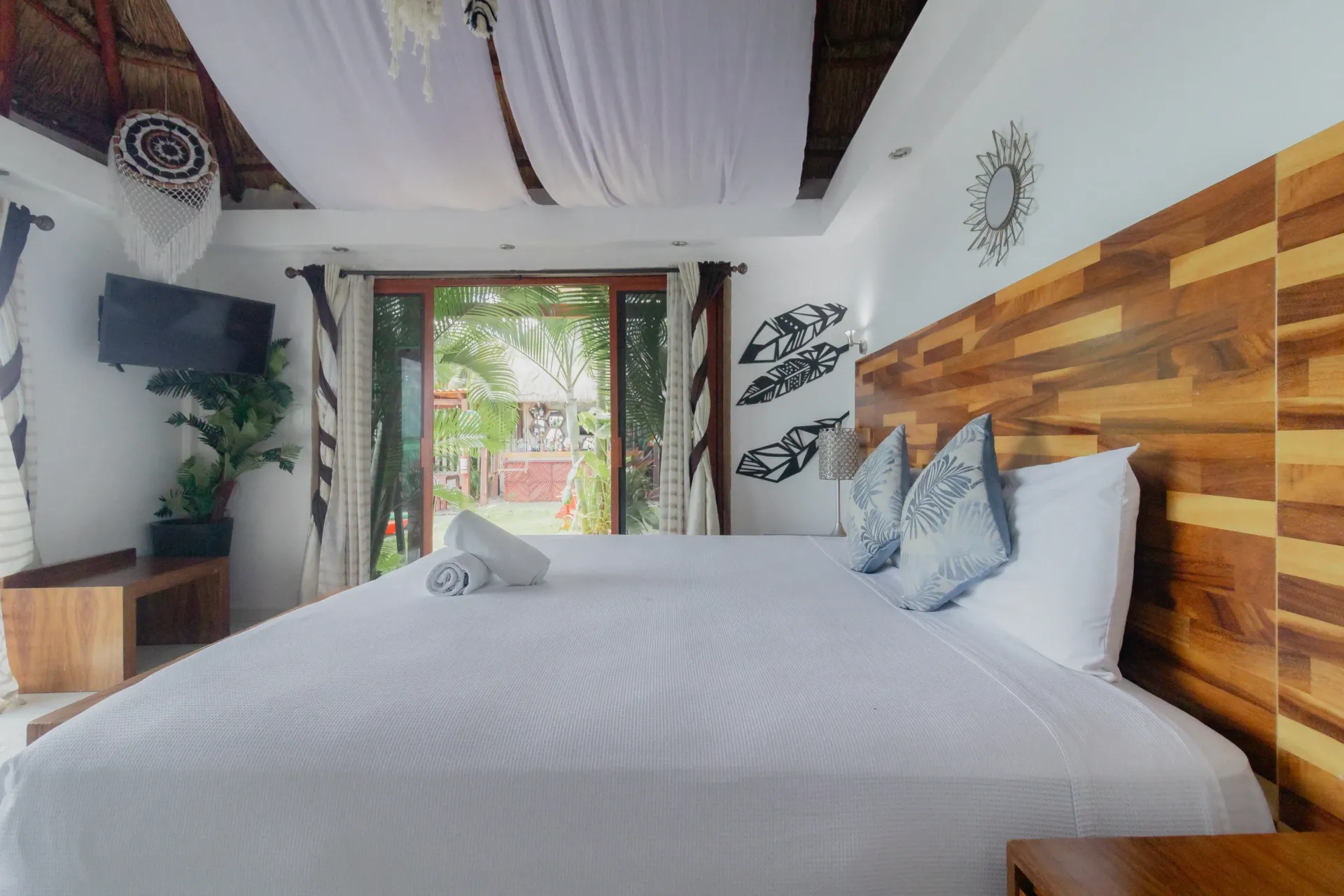 Habitación luminosa con cama blanca, cabecero de madera, plantas y puertas que dan al balcón.