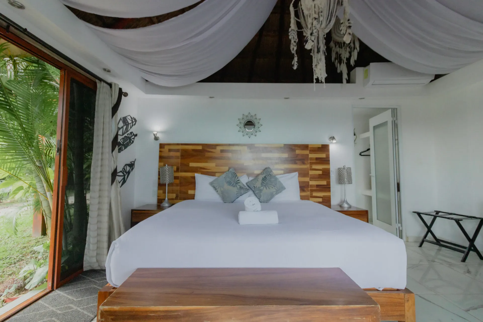 Cama blanca en una luminosa habitación tropical con dosel drapeado, detalles de madera y vista abierta al jardín.