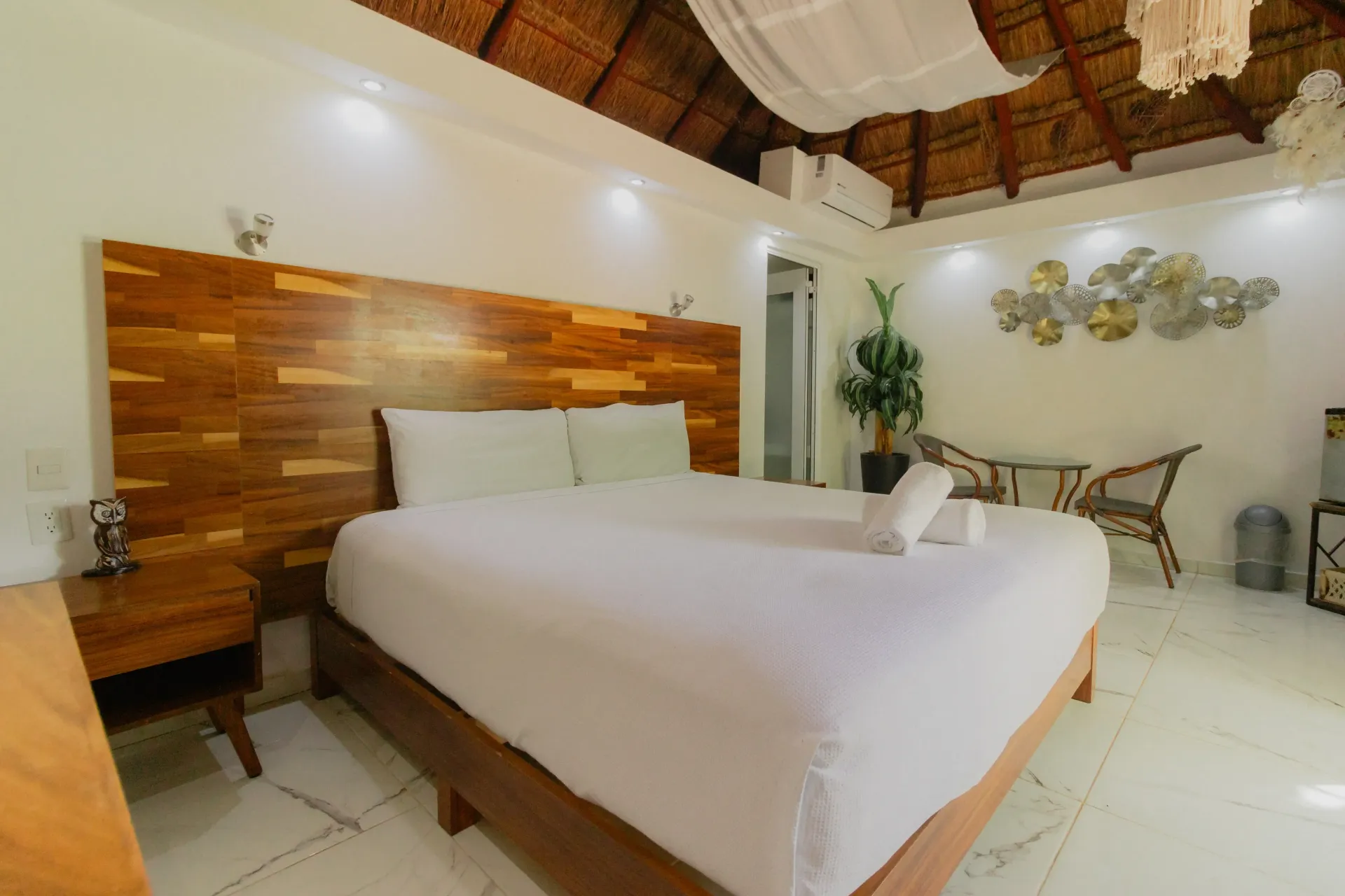 Habitación luminosa con una pared decorativa de madera, cama blanca y decoración tropical bajo un techo de paja.