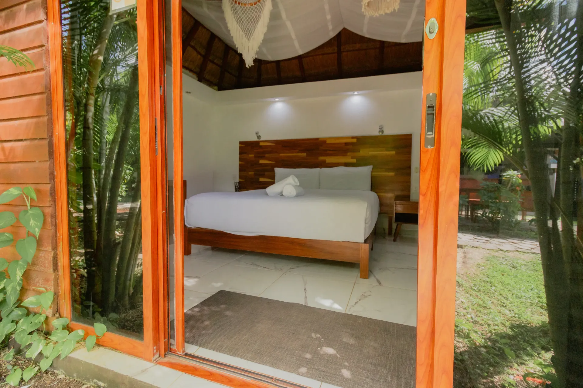 Habitación al aire libre con cama de madera, ropa de cama blanca y puertas de cristal con marcos naranjas con vistas.