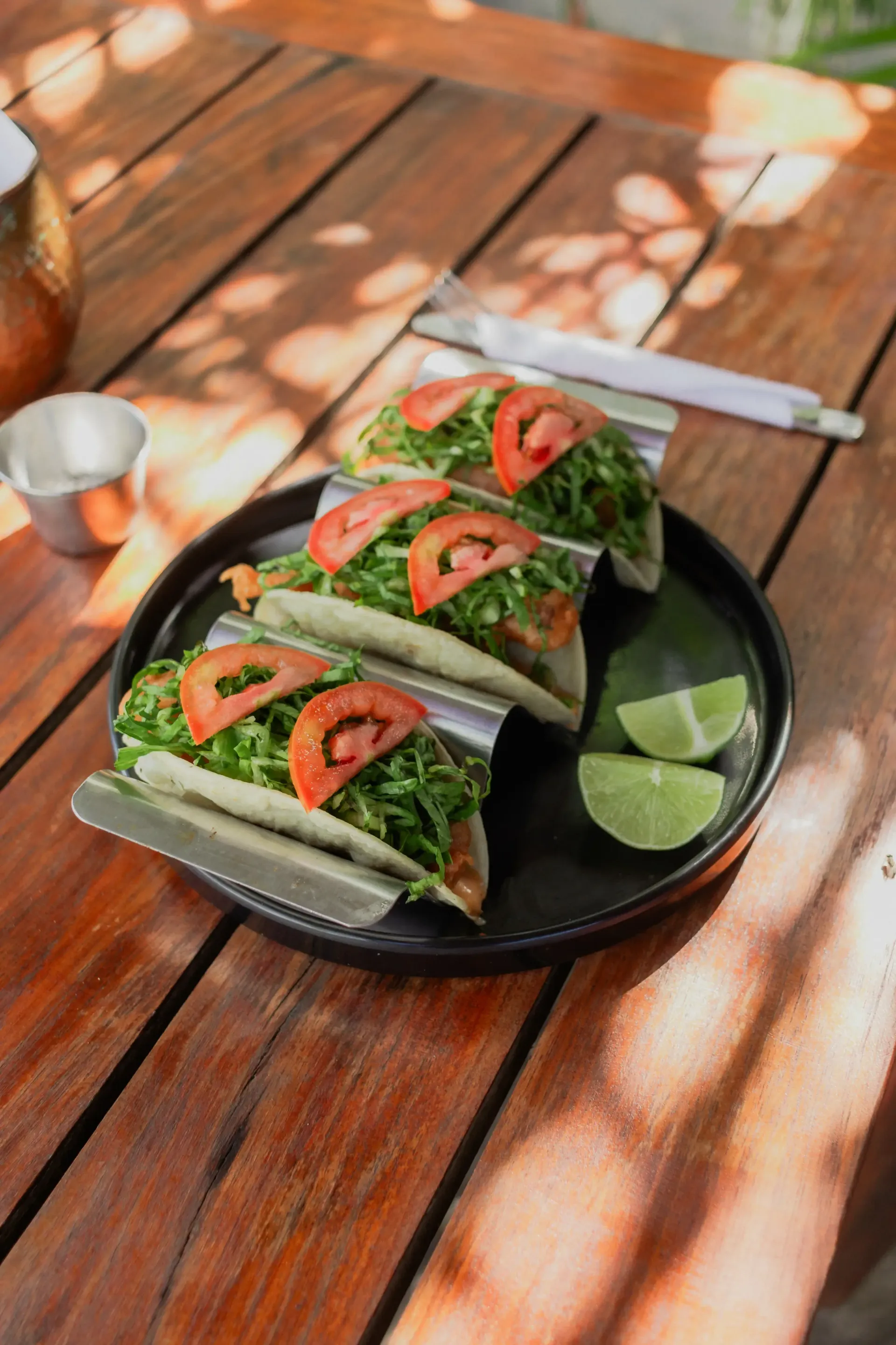 Un plato de tacos en un plato negro con rodajas de lima sobre una mesa de madera, al aire libre bajo la luz del sol.