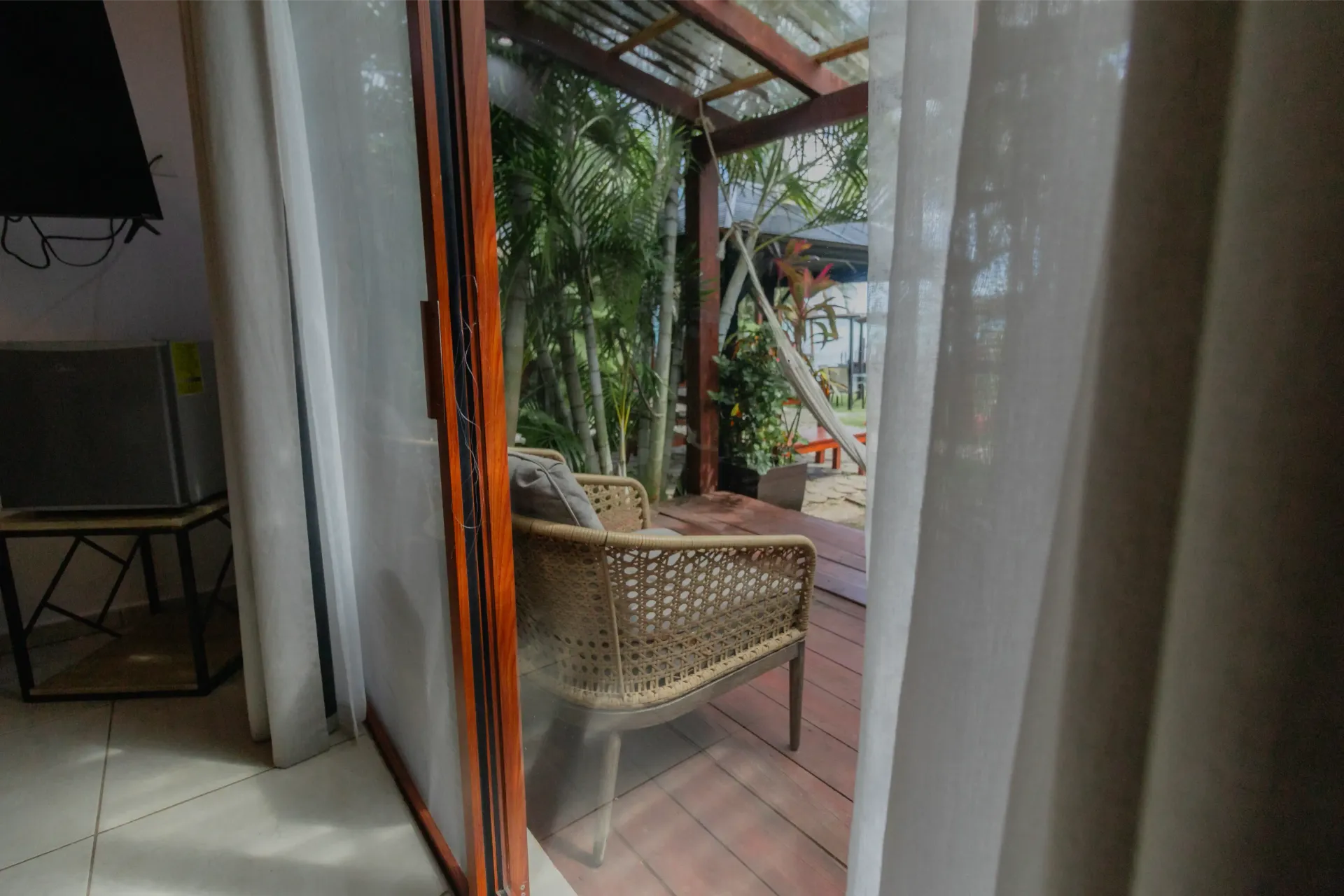 Silla de mimbre en un patio, vista a través de cortinas abiertas, con vegetación en el exterior.