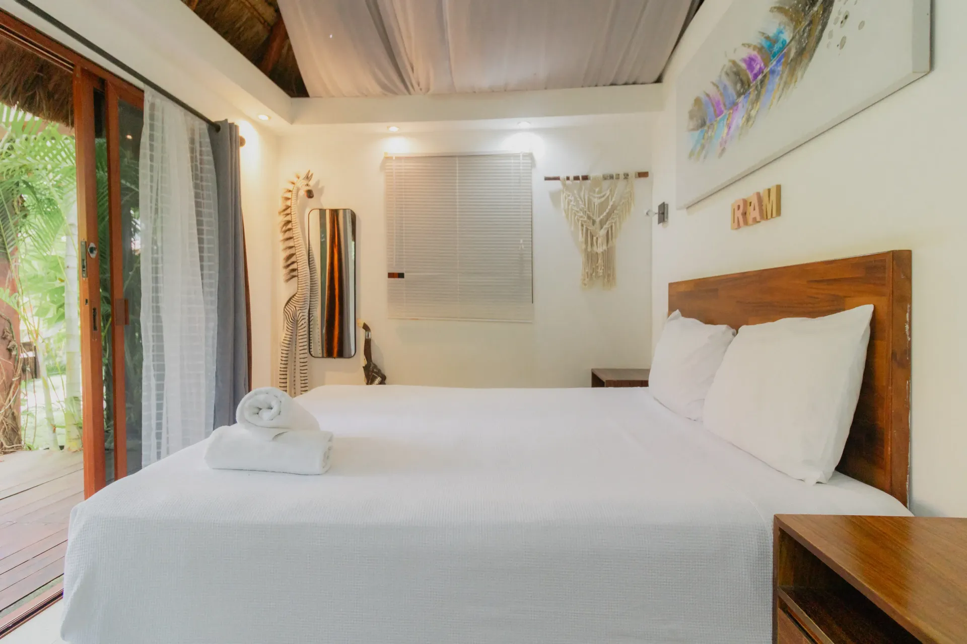 Habitación luminosa con cama blanca, cabecero de madera y pared con ventanal con vistas a la vegetación.