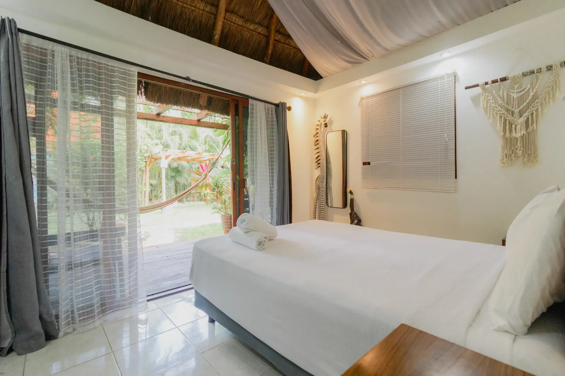 Habitación luminosa con cama blanca, cortinas transparentes, techo de madera y puertas que dan a un jardín tropical.