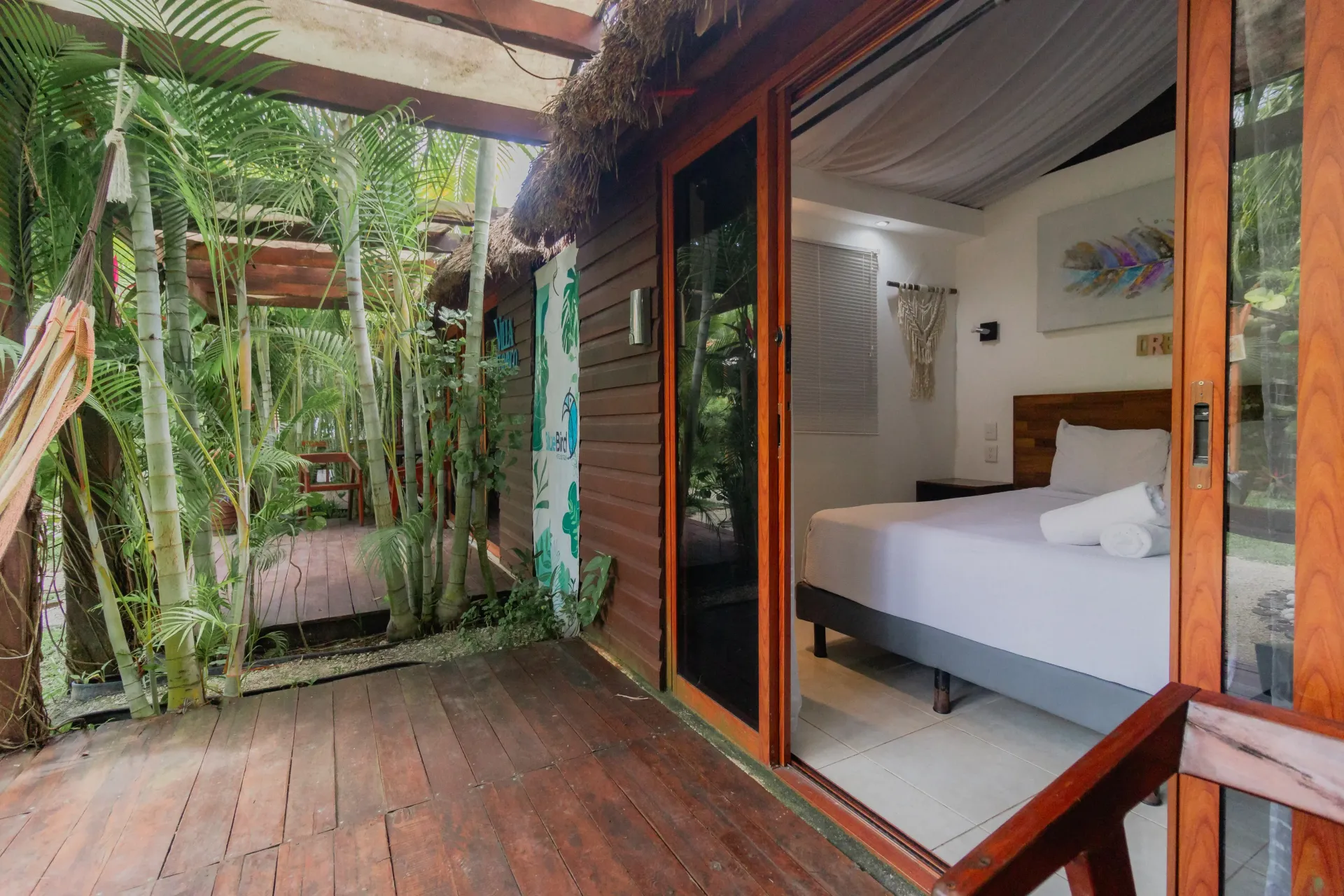 Patio de madera con plantas colgantes junto a un dormitorio con cama blanca y puertas corredizas de cristal abiertas.