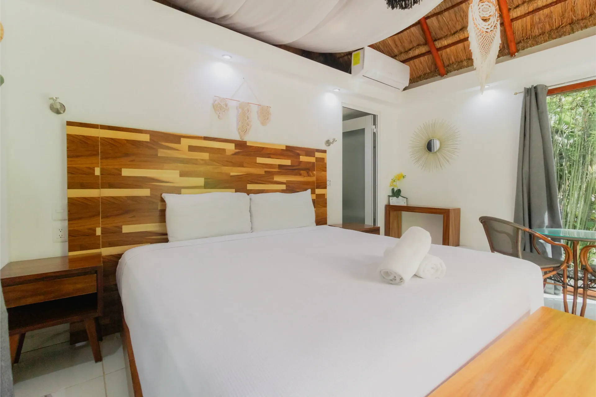 Habitación luminosa con cama blanca, cabecero de madera y vistas al balcón a través de las cortinas abiertas.