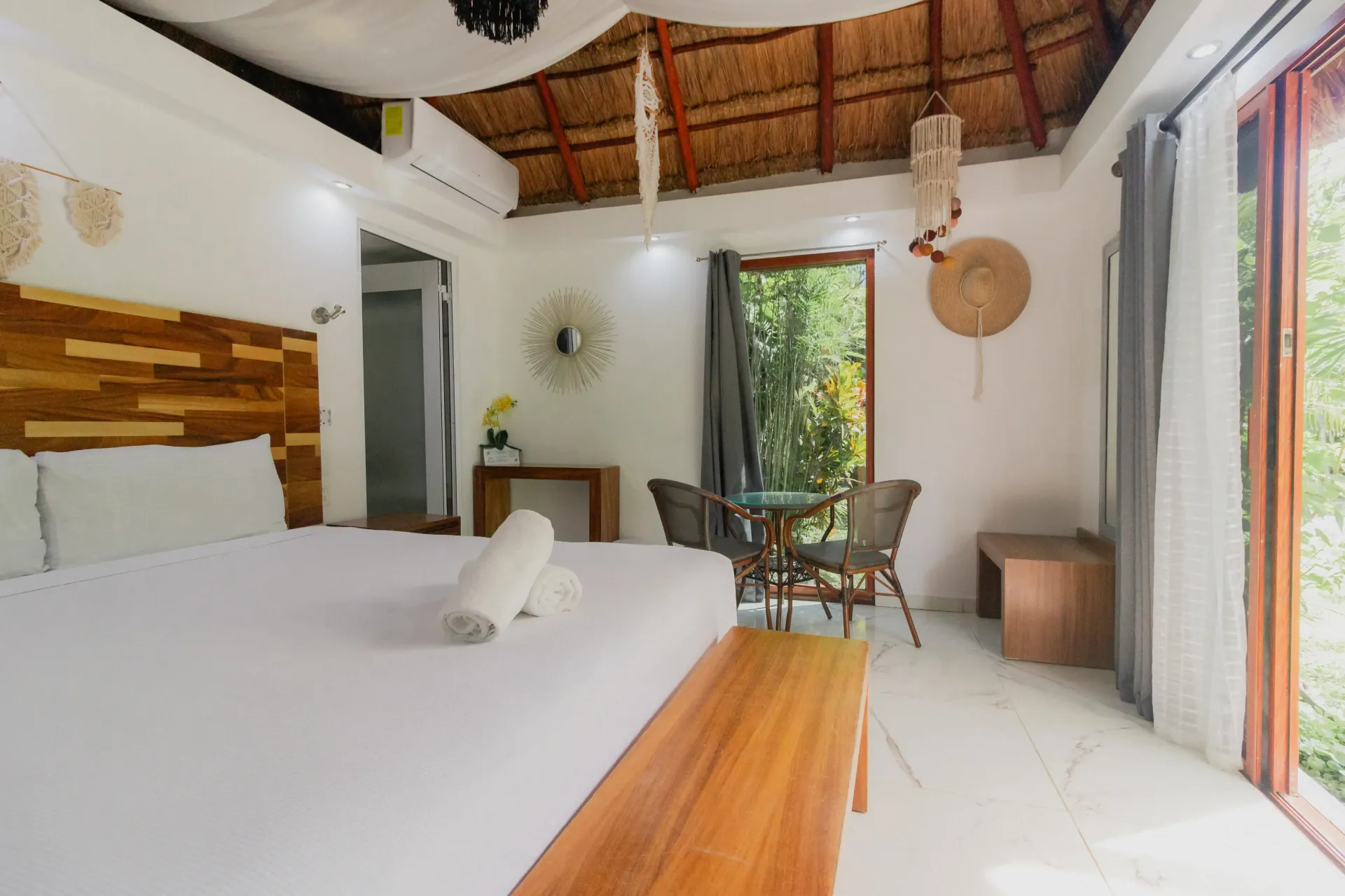 Habitación tropical luminosa con cama blanca, detalles de madera y puertas abiertas que dan a un patio ajardinado.