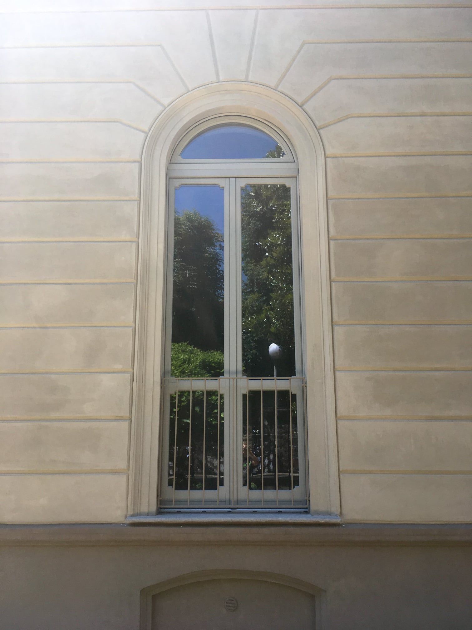 Alta finestra ad arco con cornice bianca, sbarre e una chiara vista sugli alberi, situata in un edificio beige chiaro.
