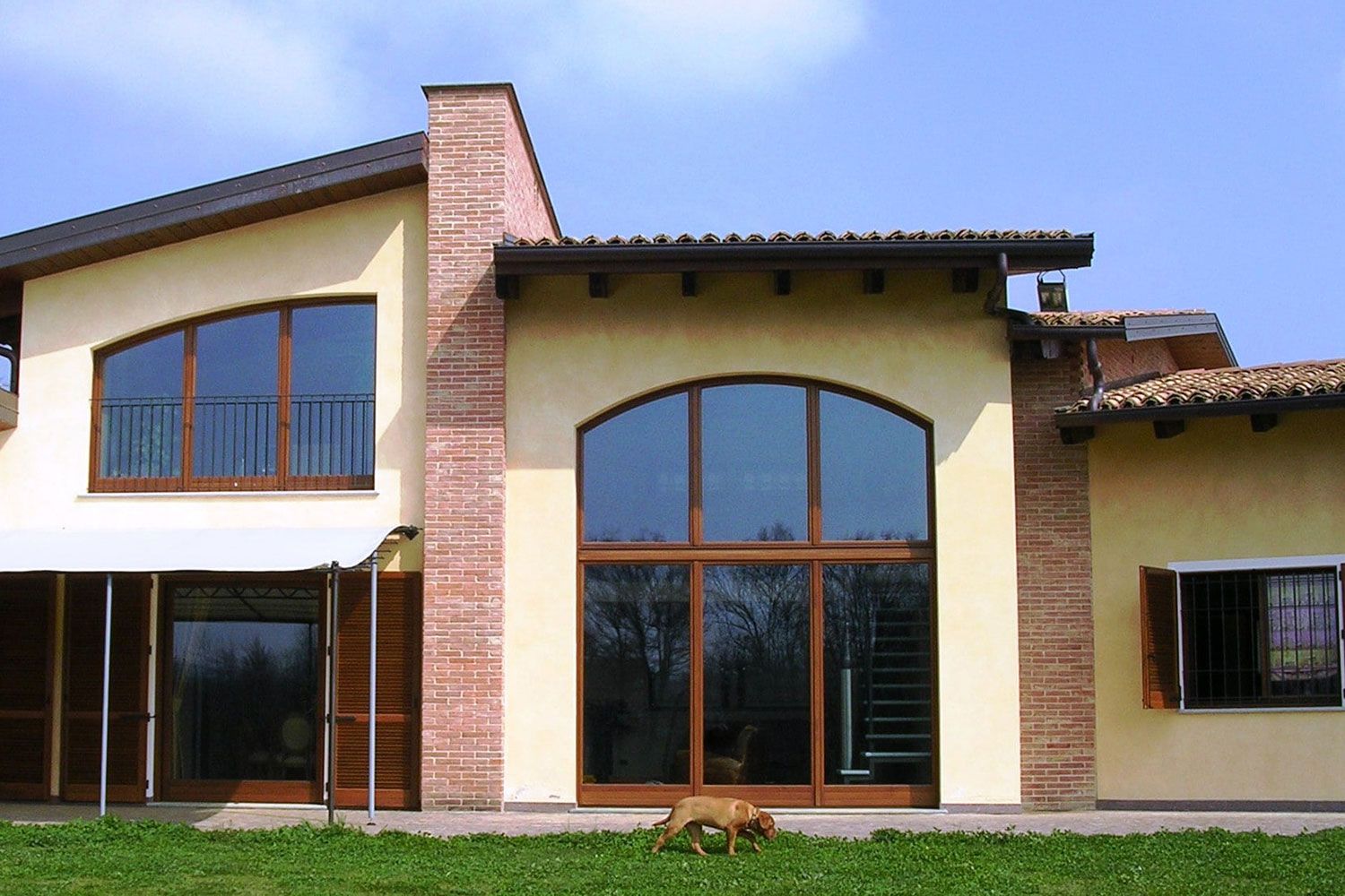 Casa beige con finestra ad arco e dettagli in mattoni, finiture marroni, cane sul prato.