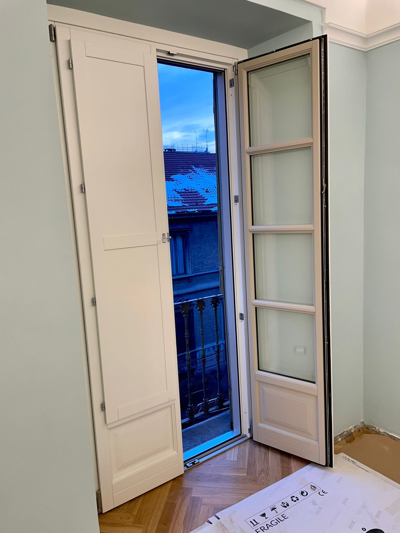 Finestra bianca aperta con persiane, con vista sul paesaggio urbano. Cielo azzurro visibile.