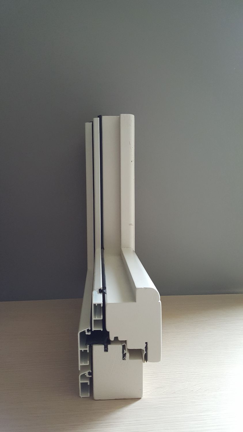 Sezione trasversale di un telaio di finestra in vinile bianco, che mostra la struttura interna, le guarnizioni e le scanalature, su uno sfondo grigio.