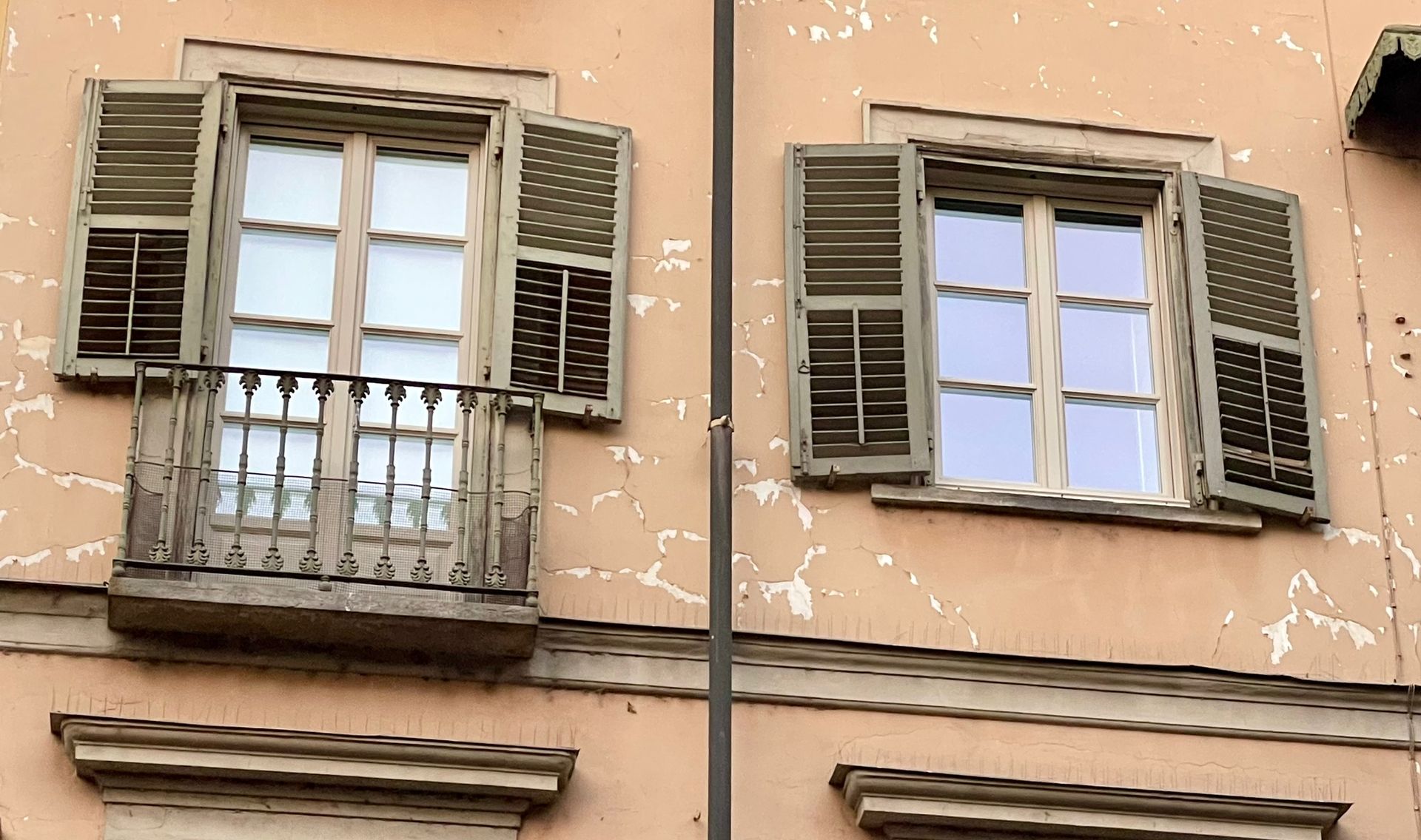 Due finestre con persiane in legno aperte su un edificio color pesca. Una ha un piccolo balcone.