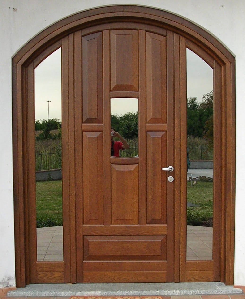Porta d'ingresso ad arco in legno con pannelli laterali in vetro, che riflettono l'esterno.