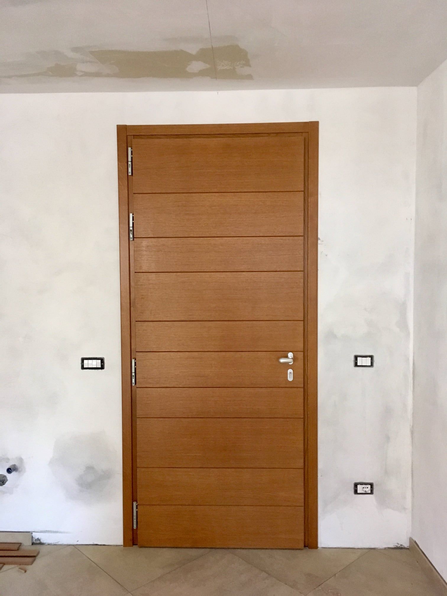 Porta in legno, marrone chiaro, inserita in una parete bianca con prese elettriche.