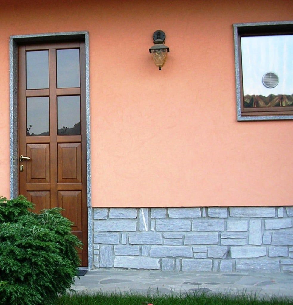 Porta e finestra marroni su un muro color pesca con una base in mattoni di pietra e un cespuglio verde.