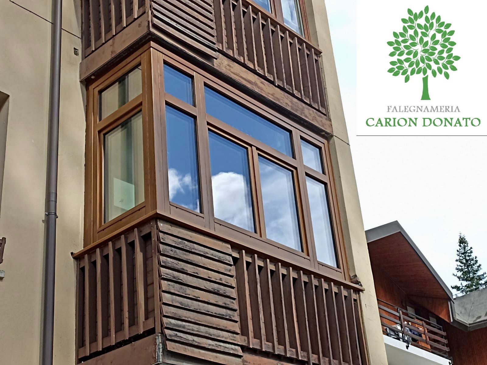 Finestra a bovindo in legno marrone con balcone, su uno sfondo di un edificio beige.