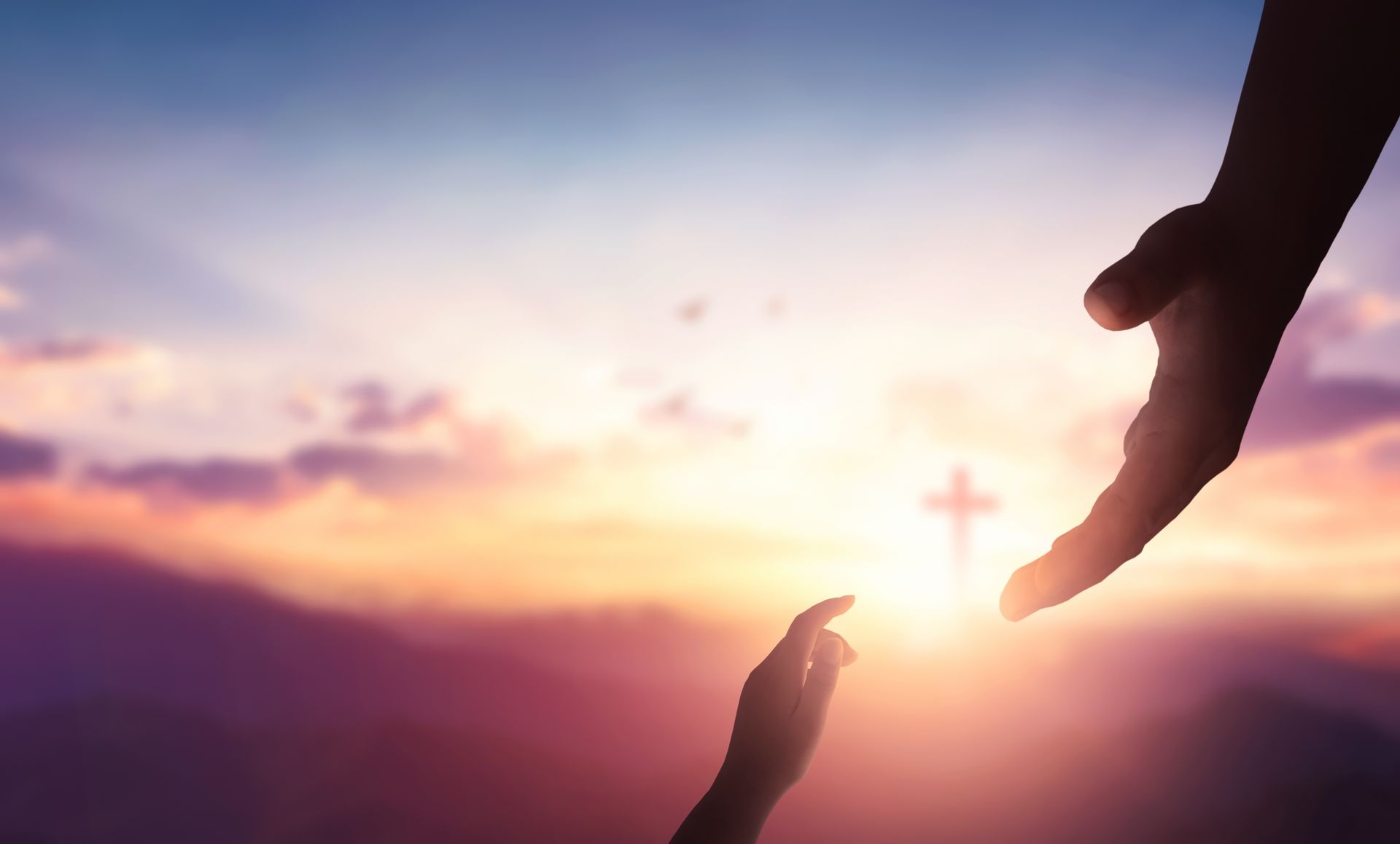 Silhouette hands reaching towards each other, backlit by a sunset and cross.