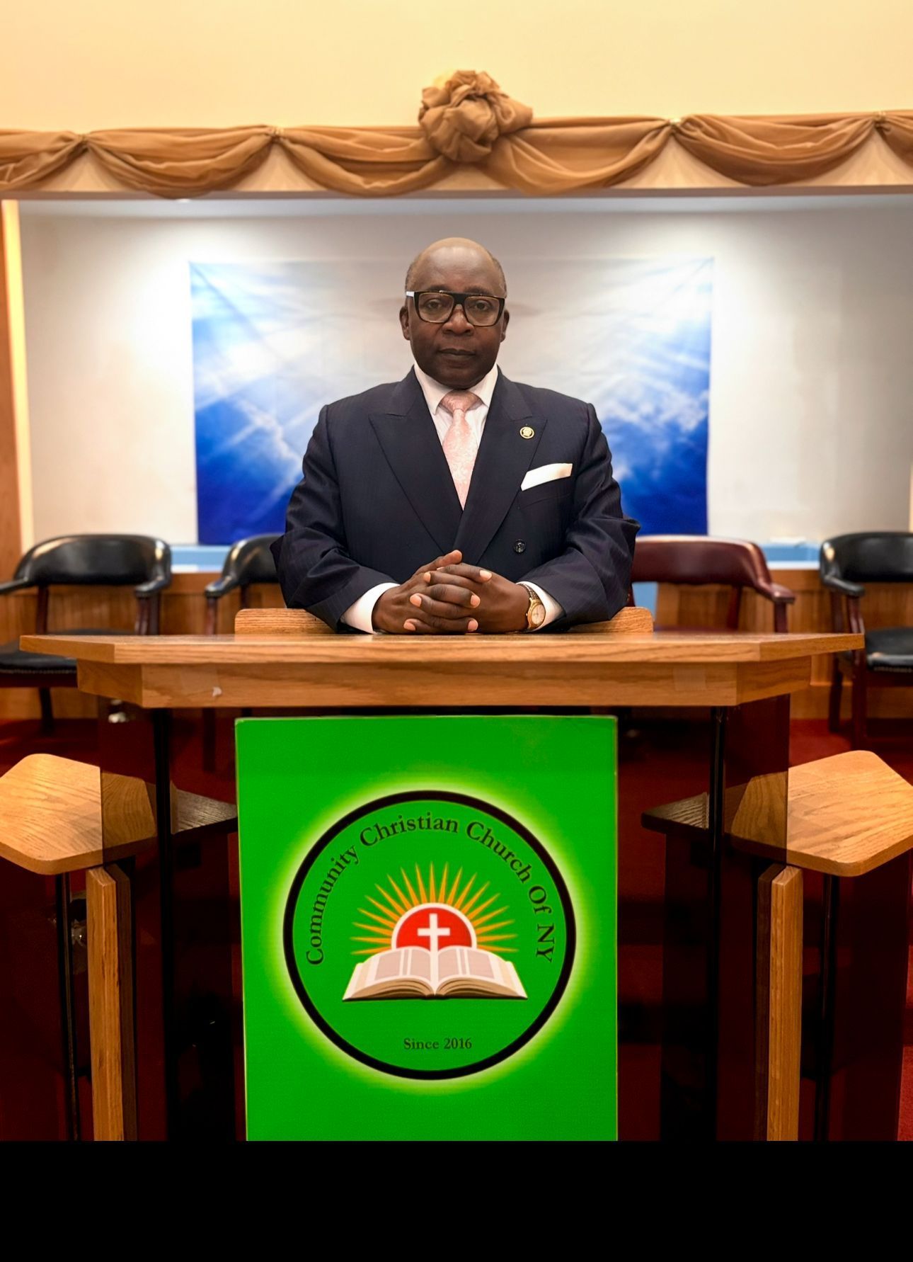 Man in suit at podium in a church; green sign with logo; blue and white background.