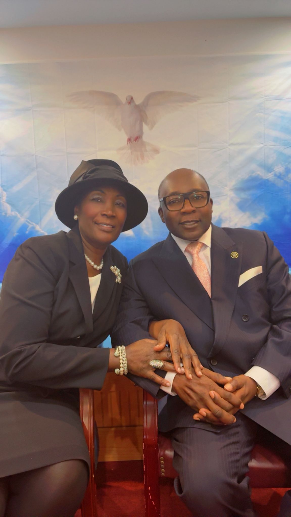 A couple sits close, hands clasped. They are in formal attire, smiling, against a cloudy backdrop with a dove.