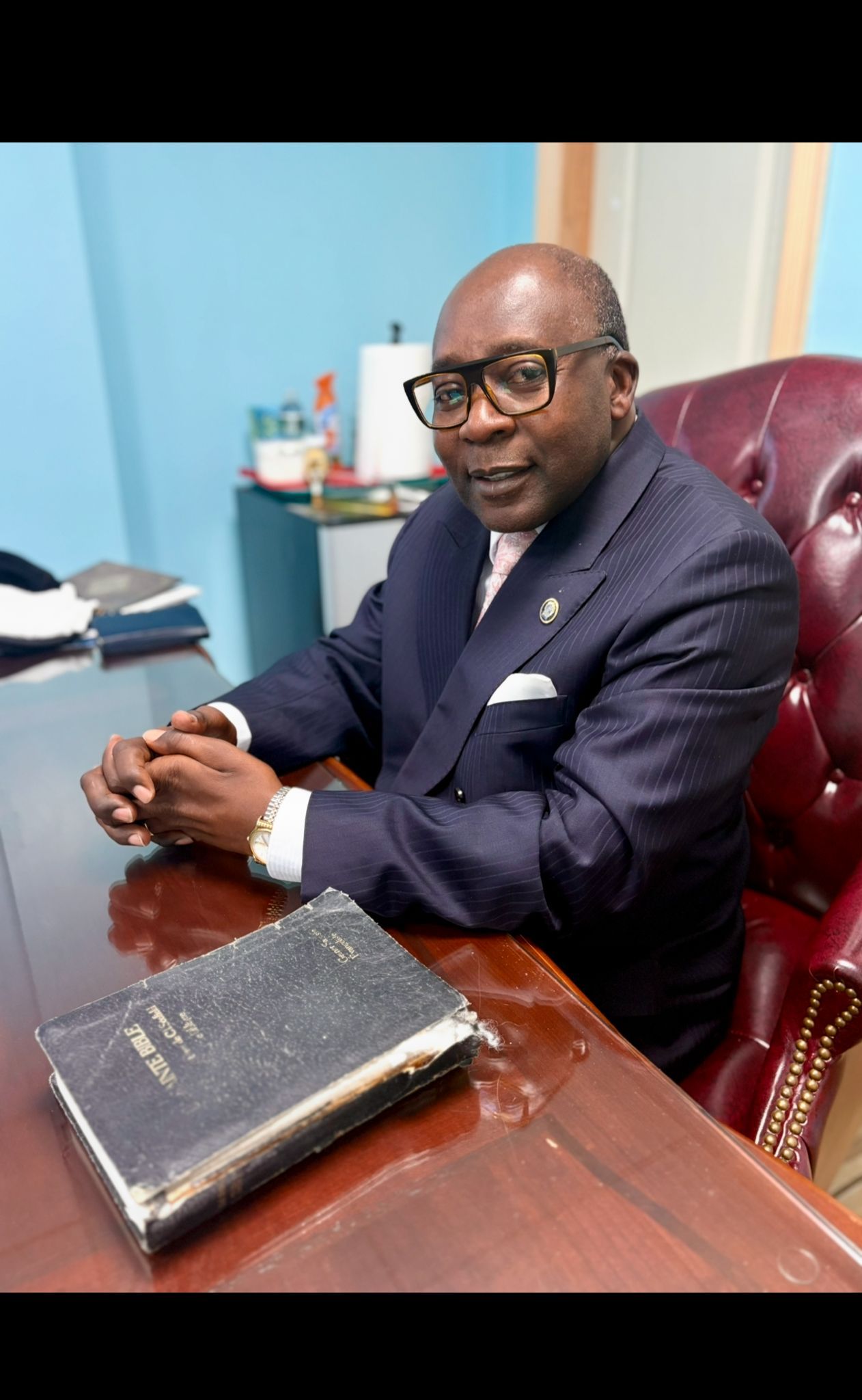 Man in suit sits at a desk, looking forward. A book lies on the desk.