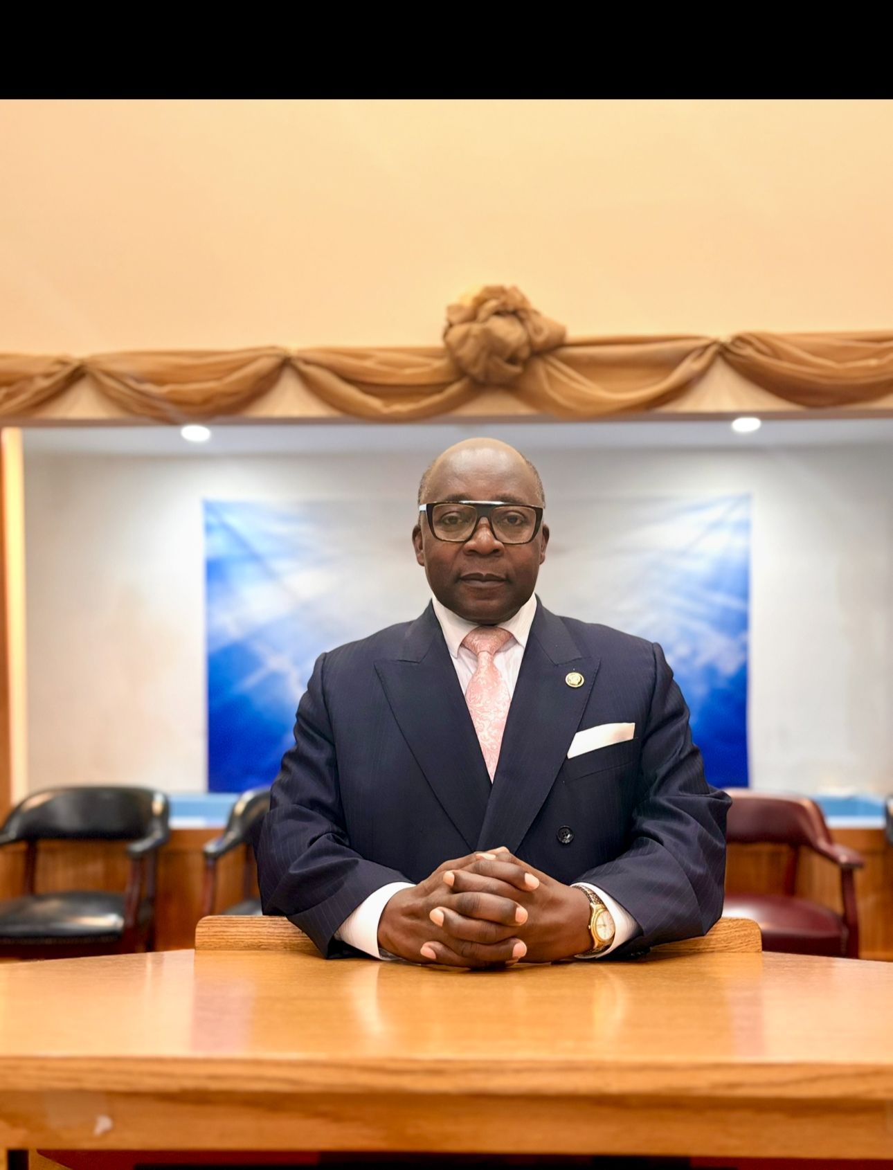Man in suit sits at a desk, arms crossed.  Behind him is a light blue backdrop and a decorative wall.