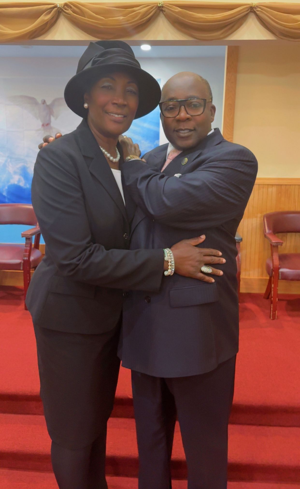 Two people embrace in a church setting. They wear suits. Red carpet and a mural backdrop.