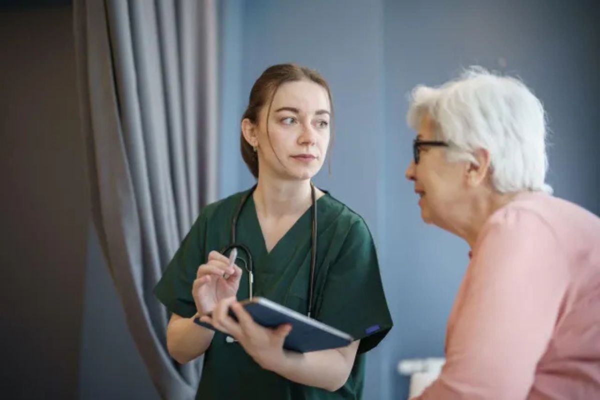 Nurse in green scrubs discussing a tablet with an older woman in a clinic room
