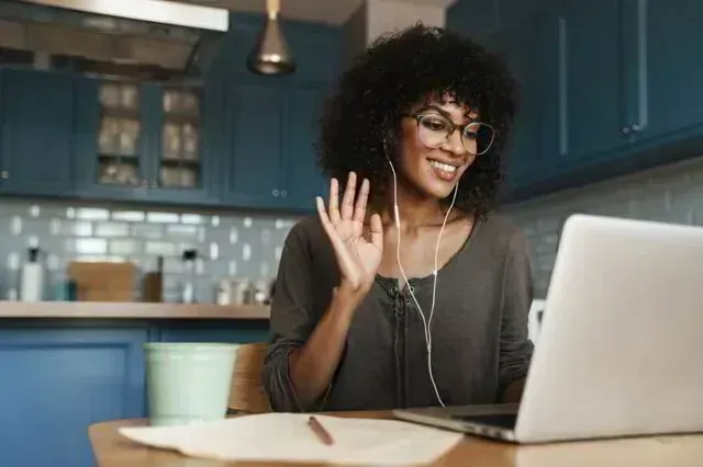 Woman smiling and waving at a laptop in a blue kitchen, with a mug and notebook on the table