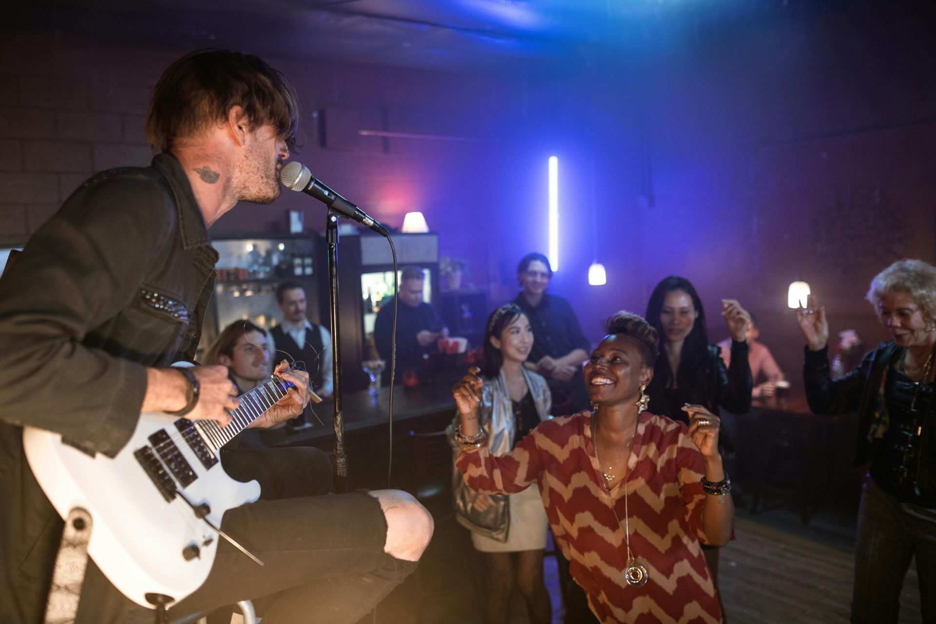 A musician performs onstage in a bar. People dance and enjoy the music in a dim-lit space.