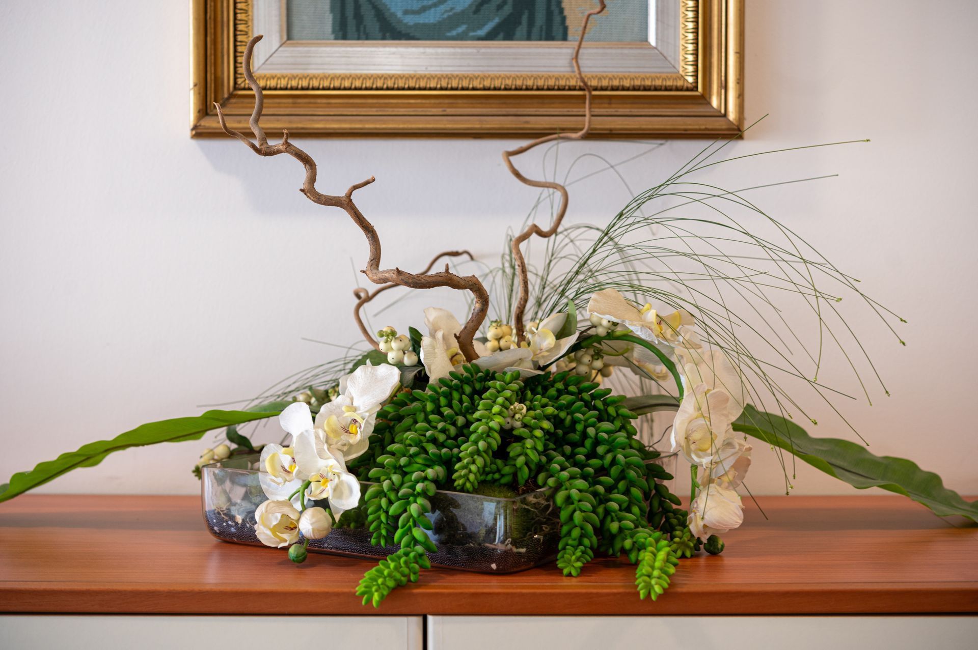 Composizione floreale con orchidee bianche, fogliame verde e rametti su un tavolo di legno sotto un quadro incorniciato.
