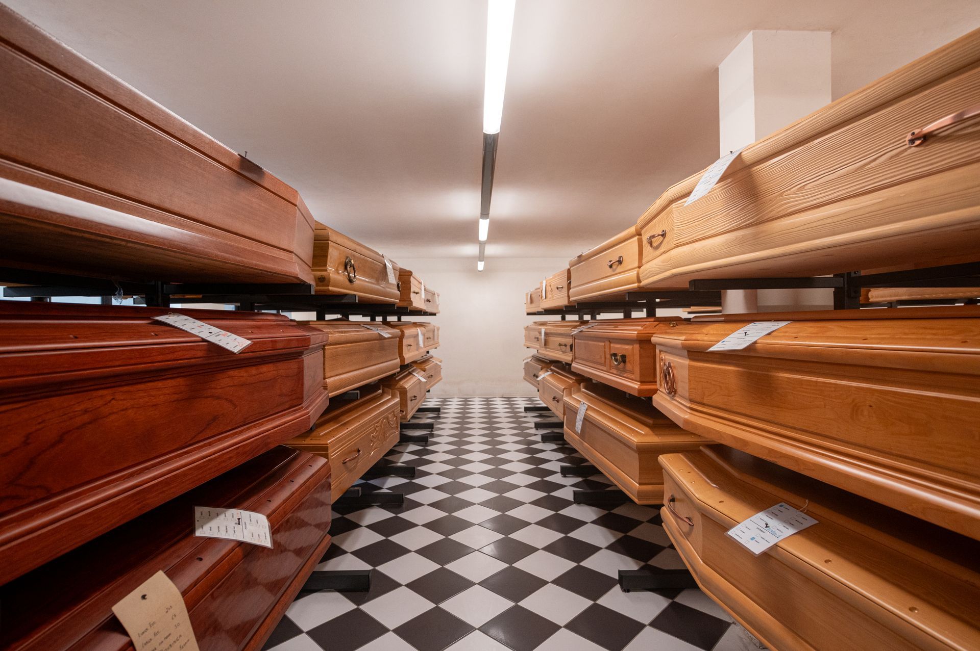 File di bare di legno nel corridoio di un'impresa di pompe funebri con pavimento a scacchi bianchi e neri.