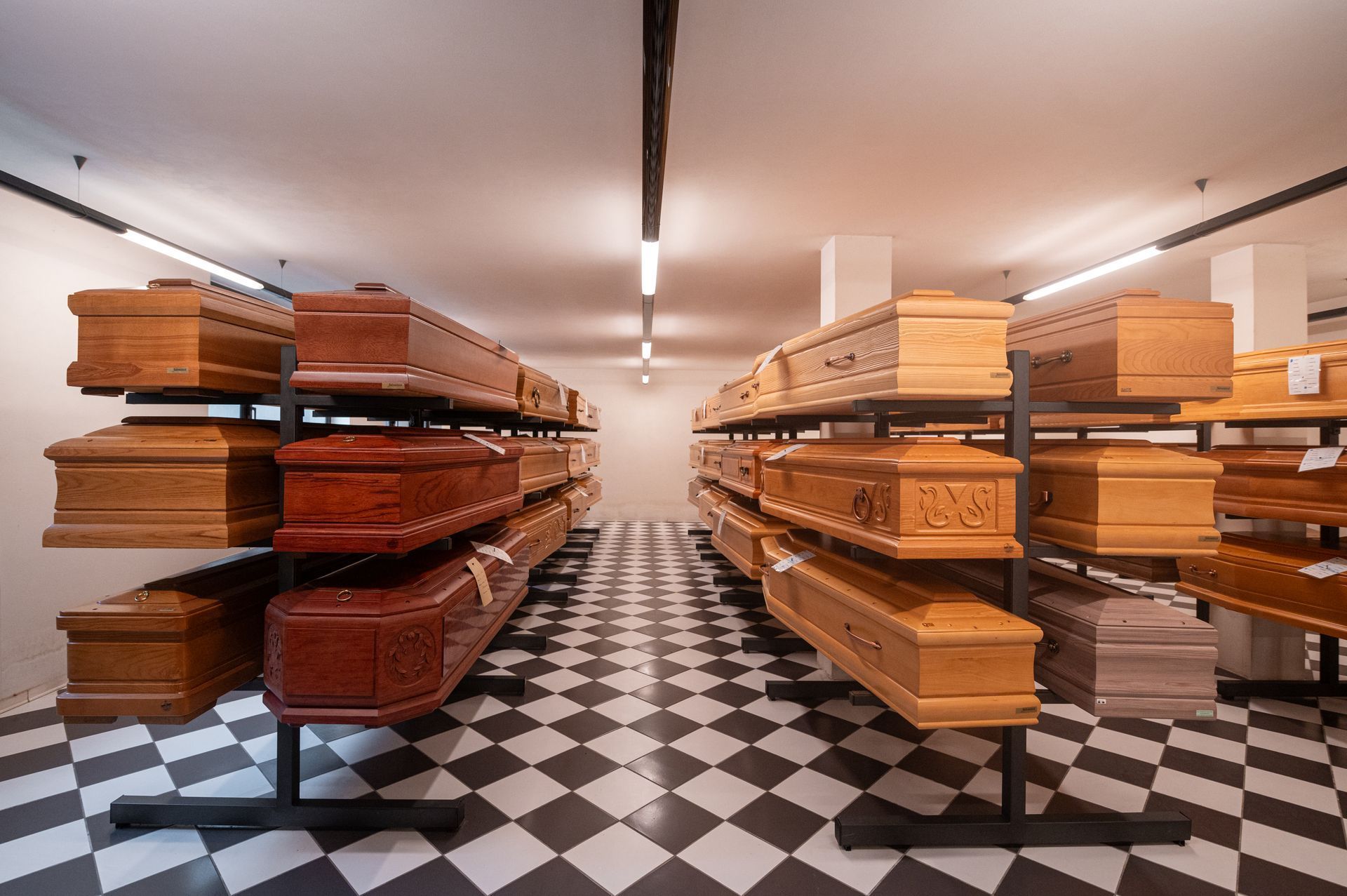 File di bare di legno su rastrelliere metalliche in un obitorio, con un pavimento a scacchi bianchi e neri.