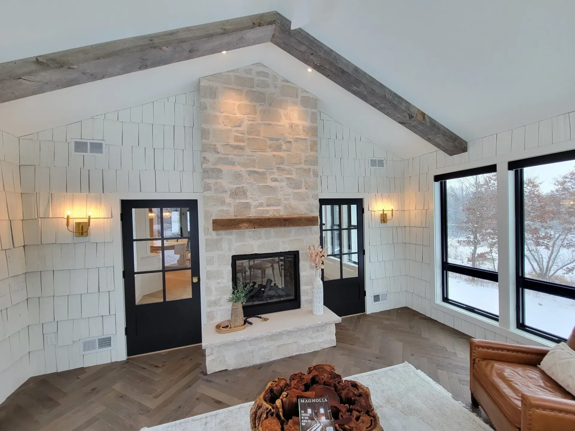 Bright living room with white stone fireplace, vaulted ceiling, dark doors, and large windows