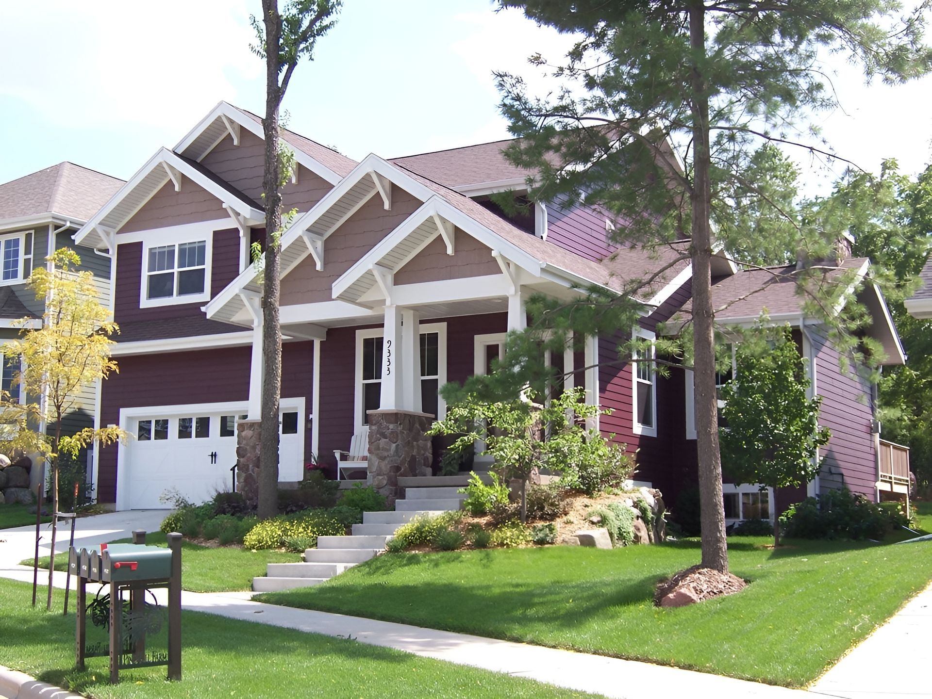 Large suburban house with a white garage, front porch, and landscaped lawn under tall trees