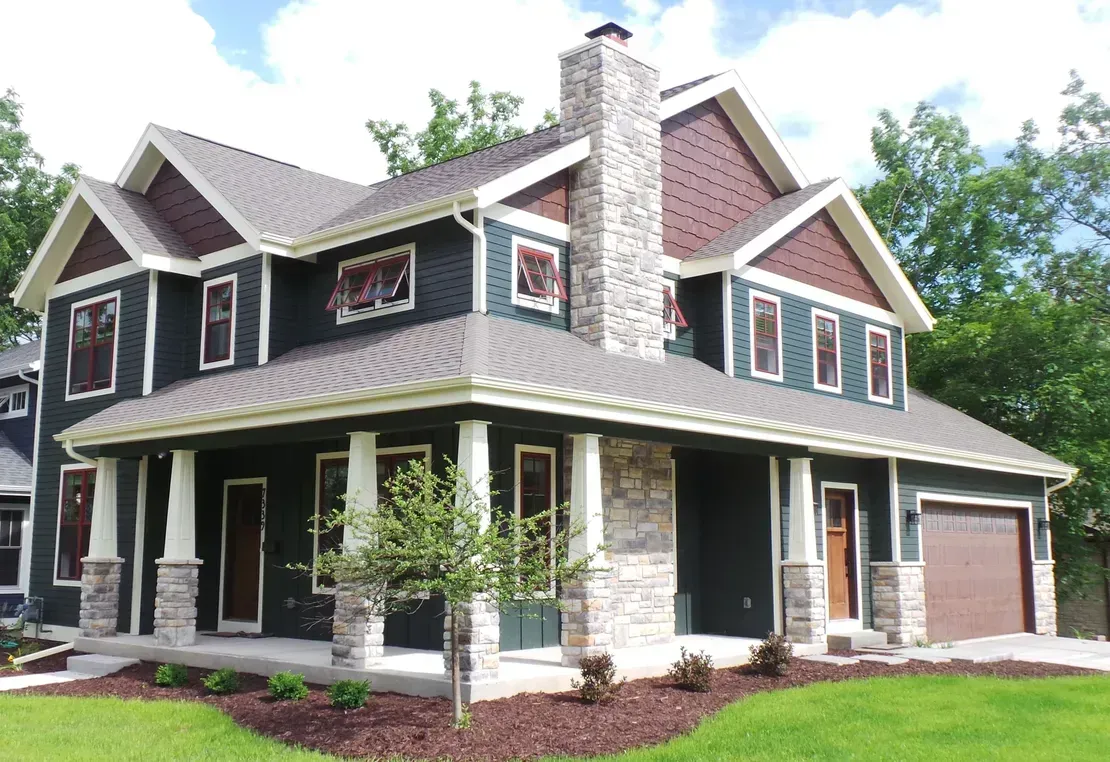 Two-story dark gray house with white trim, a front porch, stone chimney, and attached garage on a green lawn