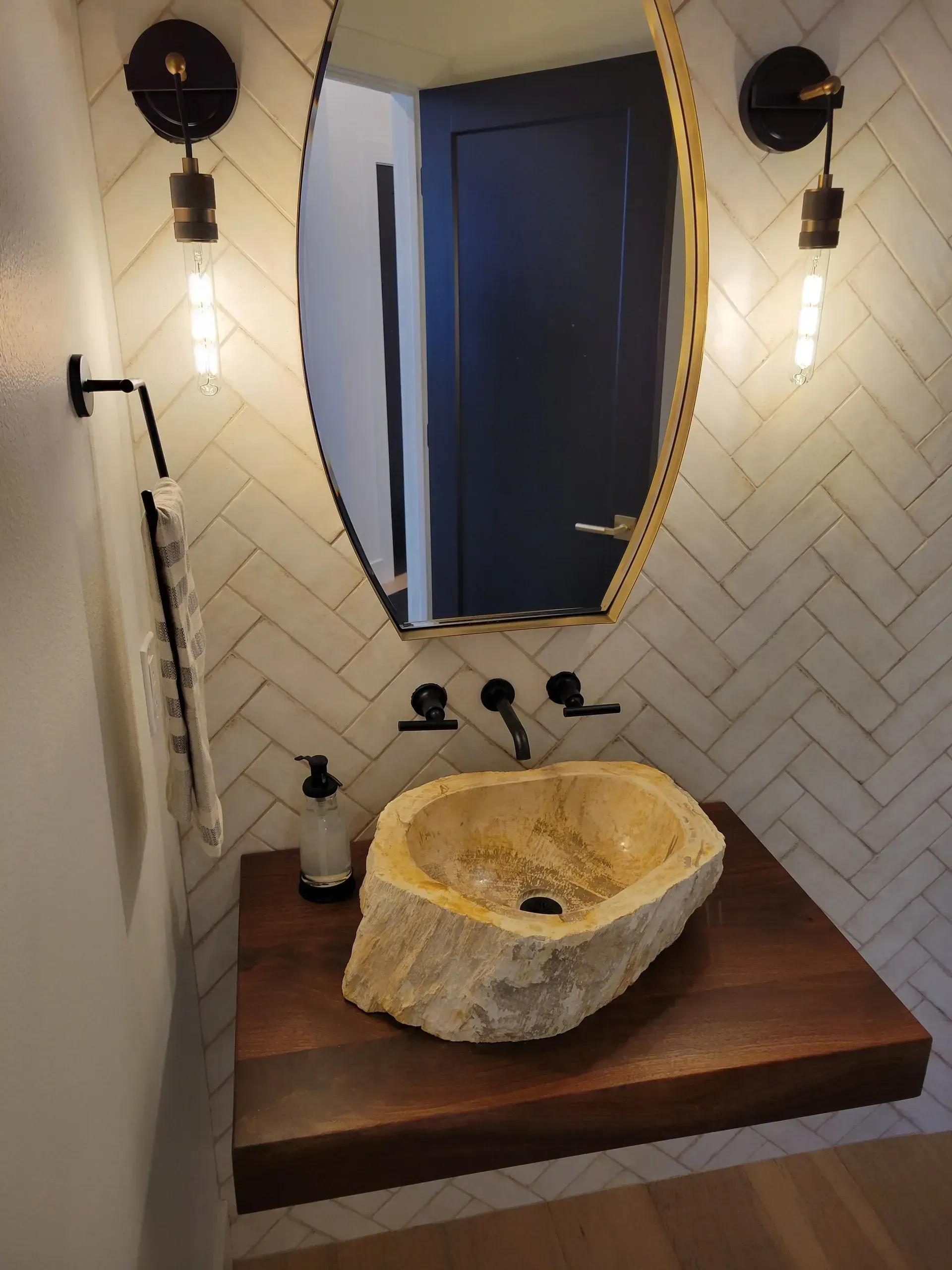 Small bathroom vanity with round stone sink, blue mirror, and wall sconces above herringbone tile.
