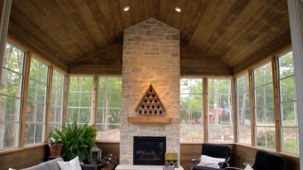 Sunroom with wood-paneled ceiling, brick fireplace, and large windows overlooking trees