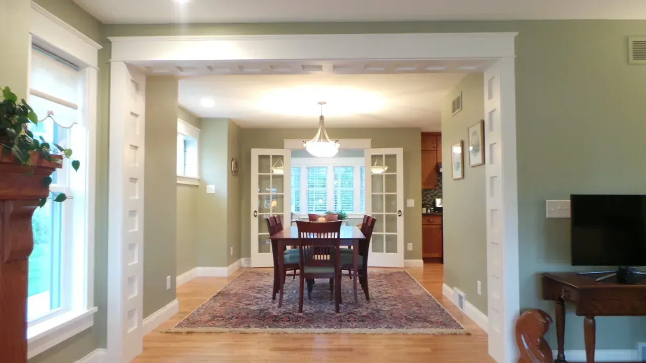 Dining room with wooden table, chairs, chandelier, and patterned rug, viewed through a wide entryway