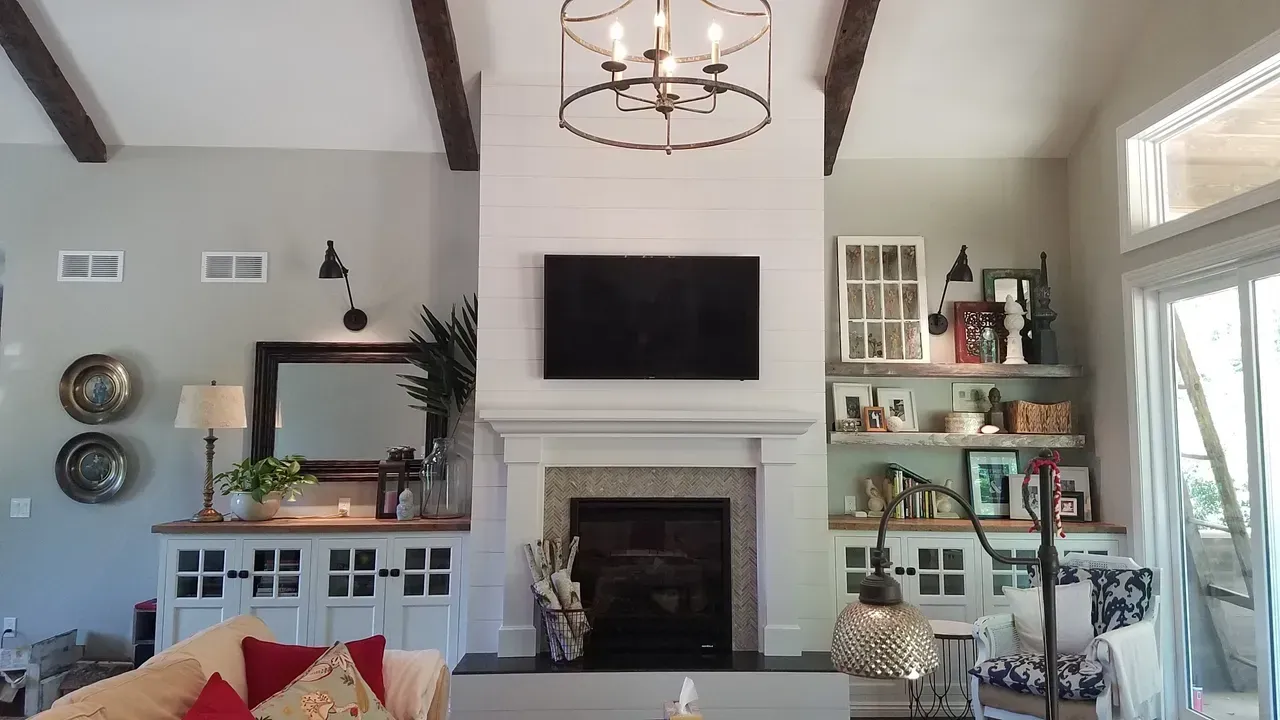Bright living room with fireplace, wall-mounted TV, shelving, mirror, and large windows.