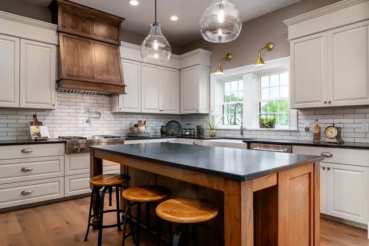 Bright kitchen with white cabinets, dark island, pendant lights, and wooden stools under a large hood.