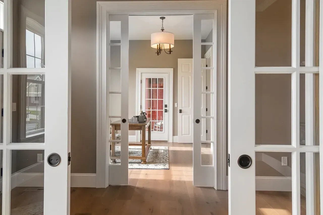 Bright hallway with white French doors, leading to a sunlit room with a hanging light and rug.