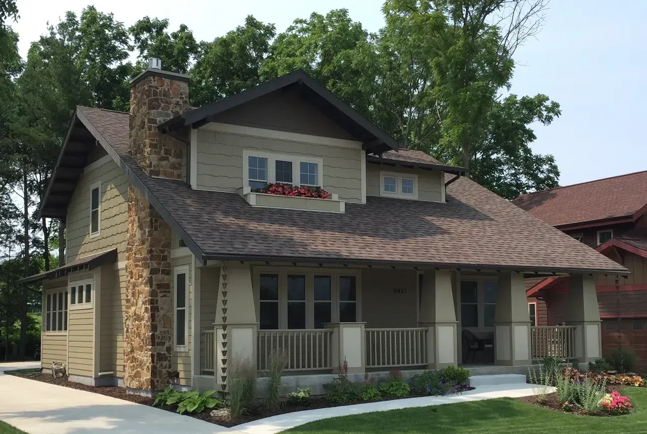 Two-story beige house with stone chimney, front porch, and landscaped yard