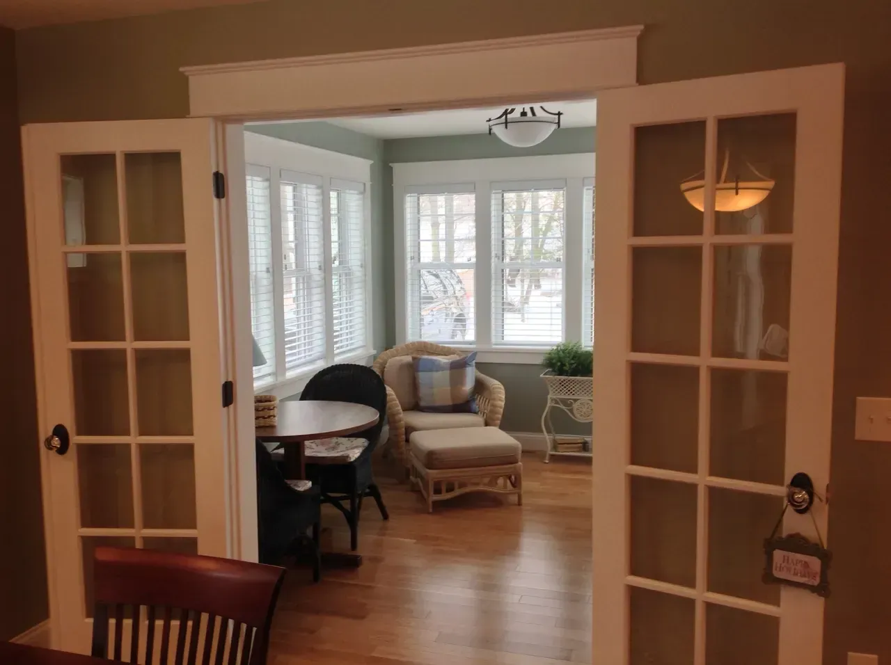 Open French doors to a bright sunroom with wicker chair, round table, and bay windows.