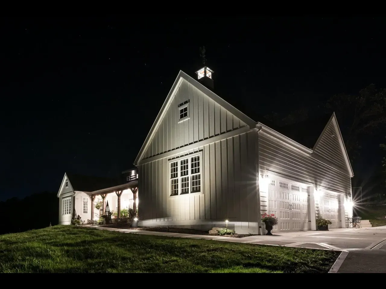 Lit house at night with white siding, gabled roof, and illuminated garage on a grassy lawn