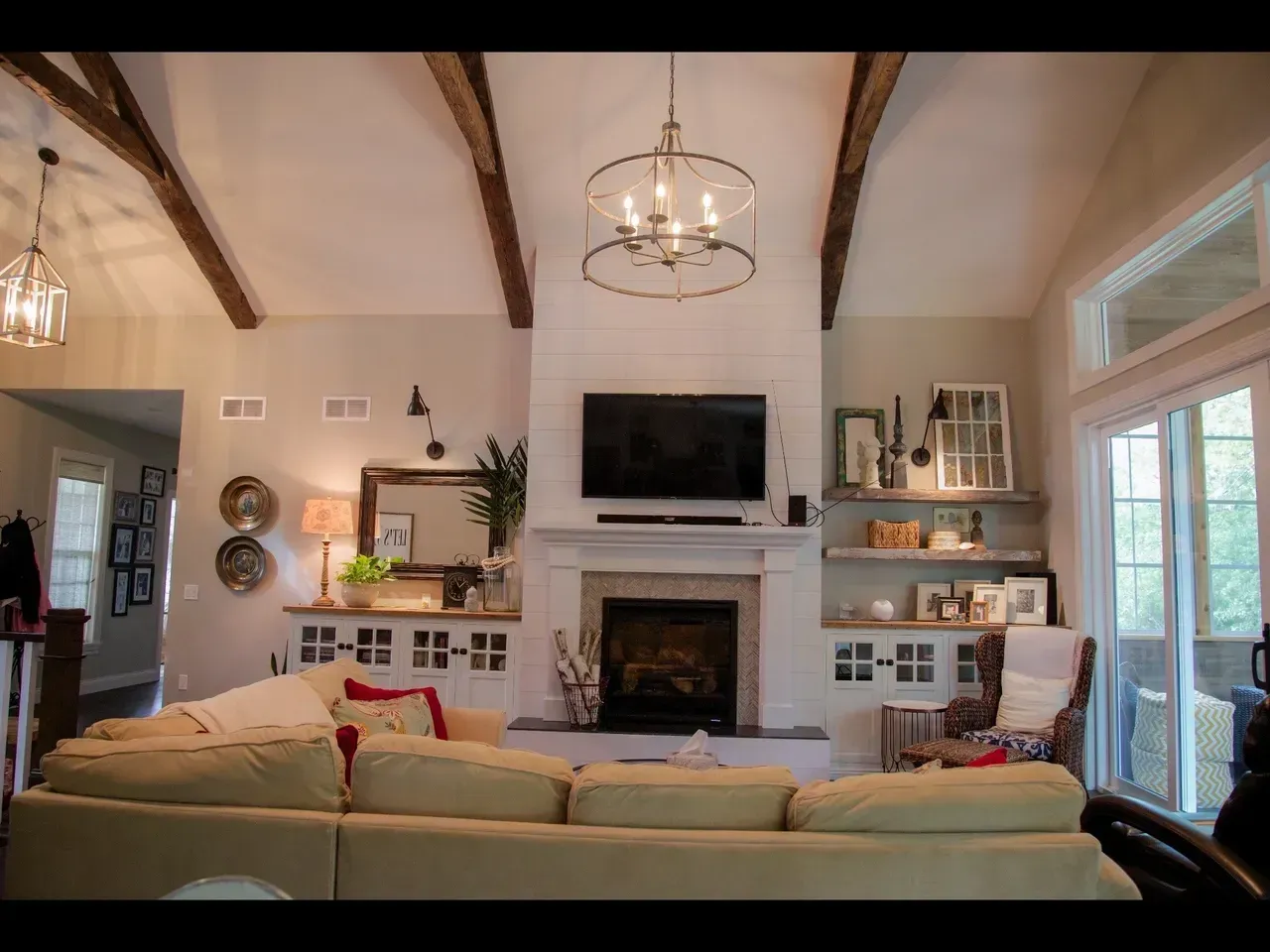 Cozy living room with vaulted wood beams, fireplace, wall-mounted TV, beige sofa, and glass doors to the patio