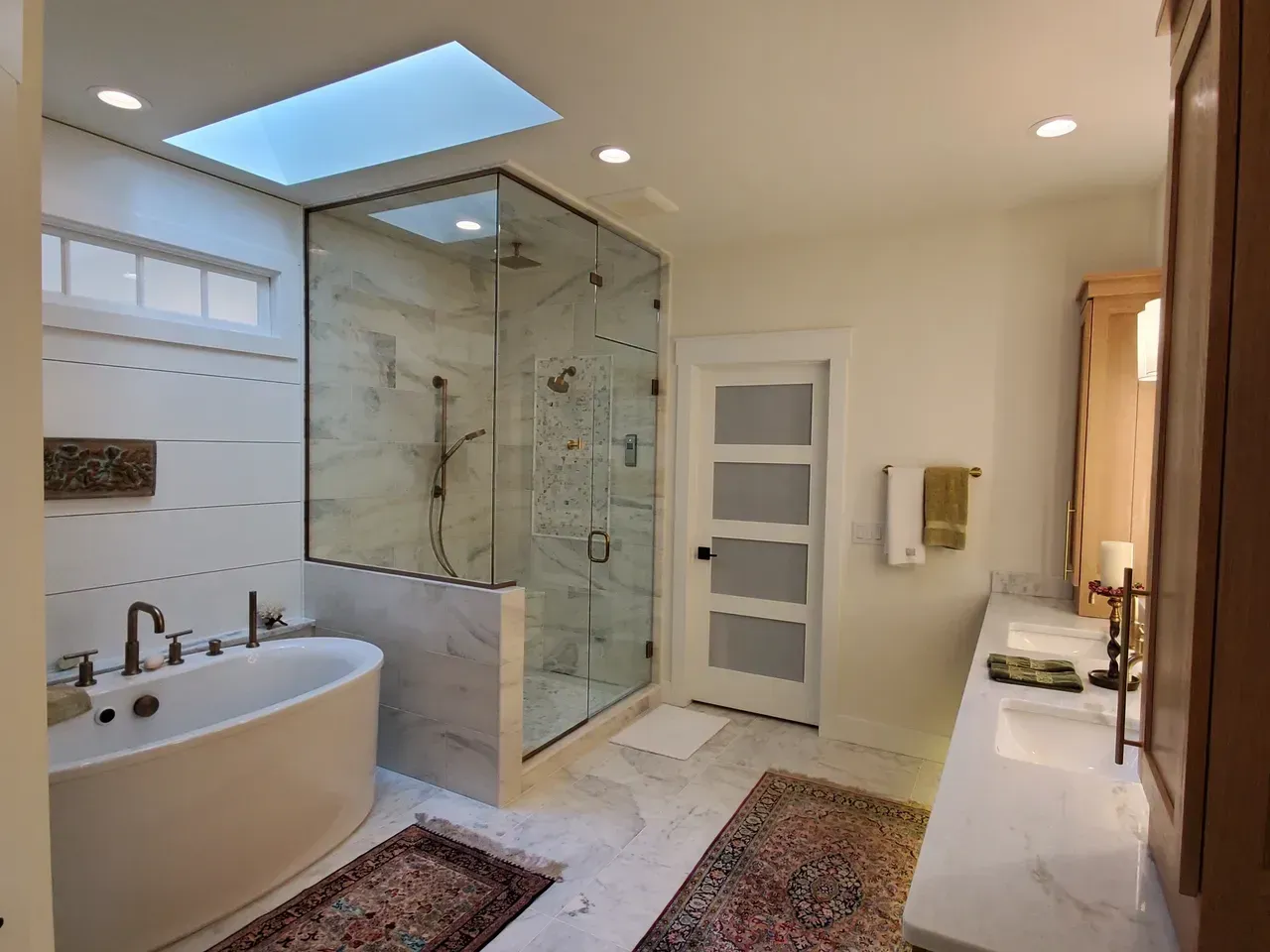 Modern bathroom with freestanding tub, glass shower, double vanity, skylight, and patterned rugs