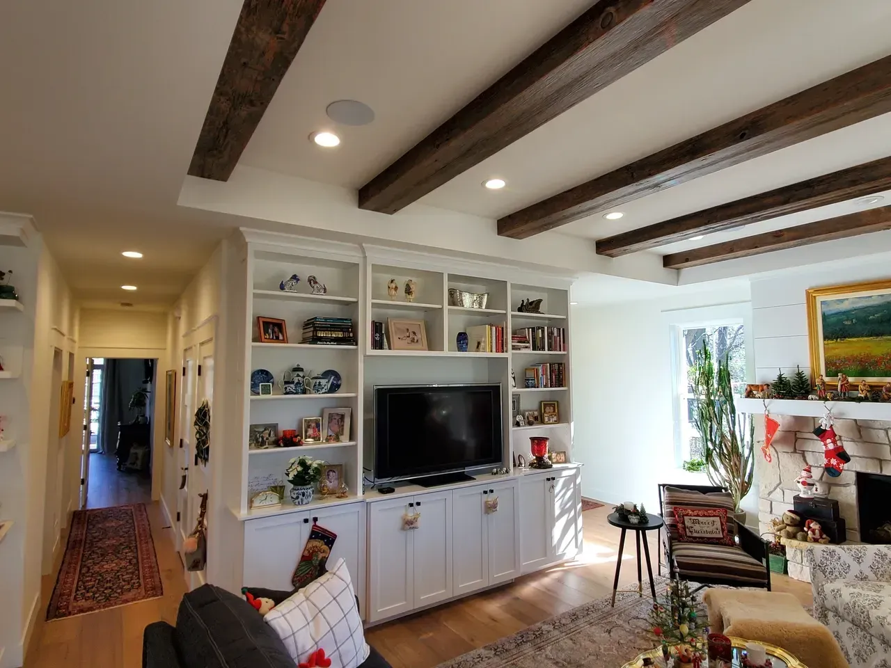 Bright living room with white built-in shelves, TV, wood ceiling beams, fireplace, and large window seating area