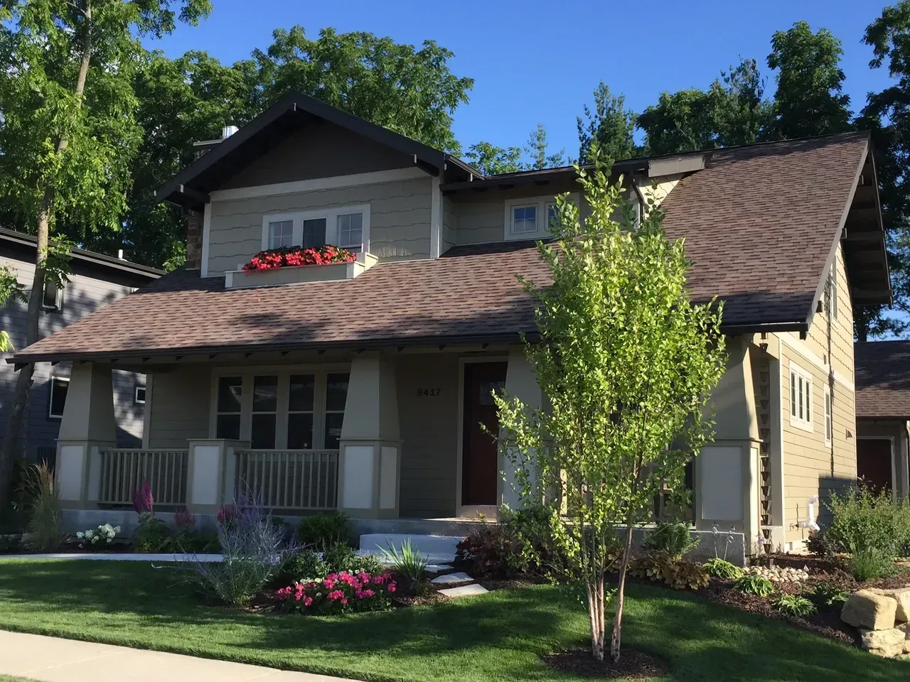 Two-story house with gray siding, brown roof, front porch, and flower garden in a sunny yard