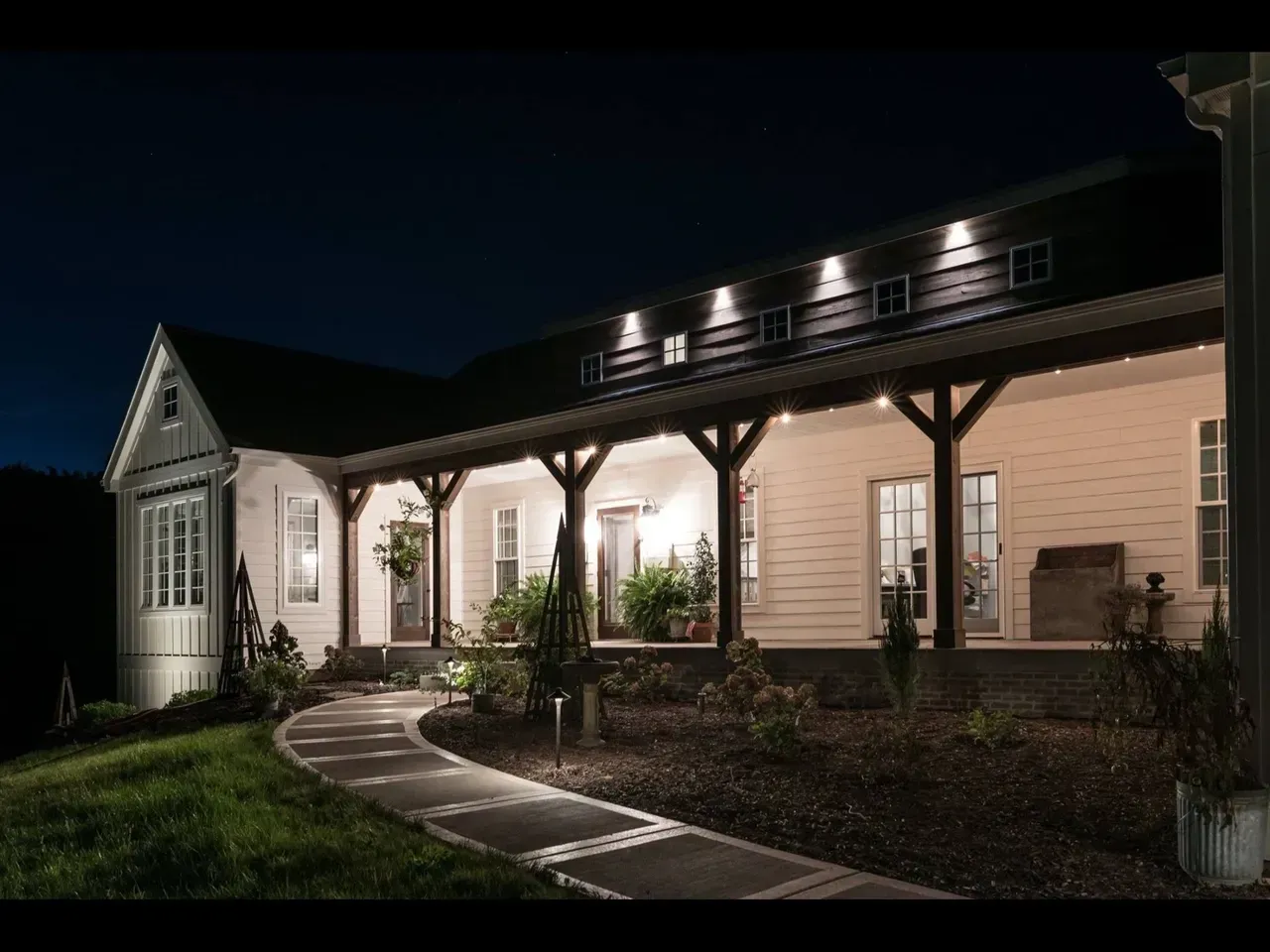 Well-lit white farmhouse porch at night with a curved stone walkway and landscaped garden
